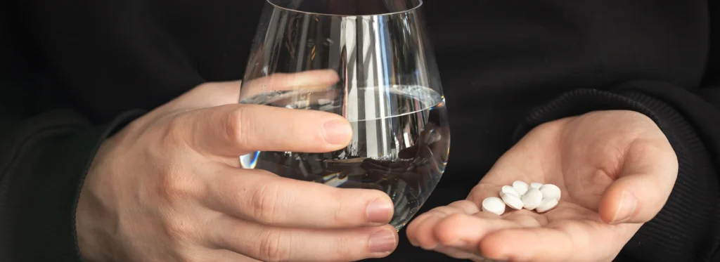 Persona sosteniendo varias pastillas en una mano y vaso de agua en la otra mano. 